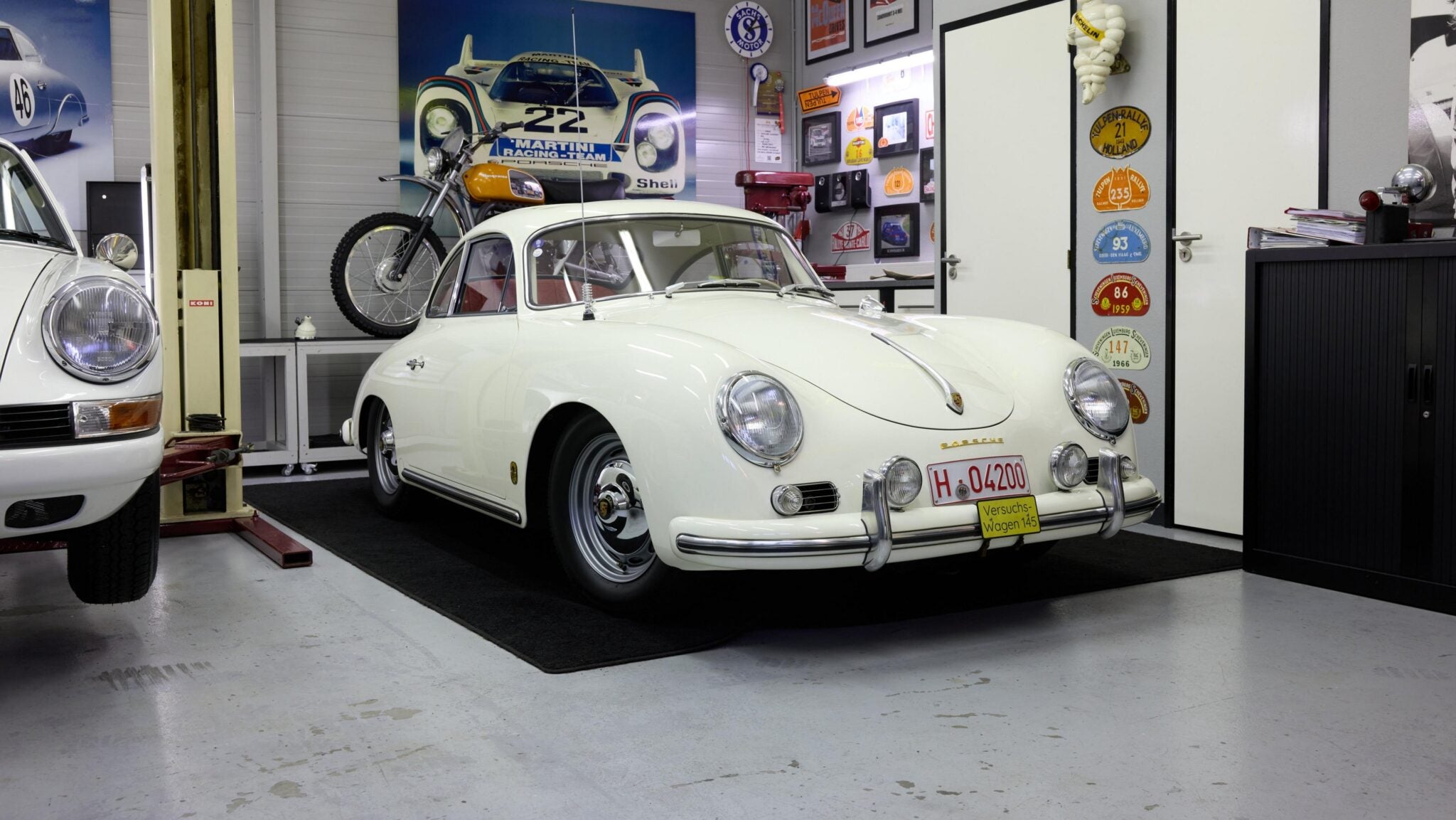 A white vintage Porsche 356 coupe is parked indoors on a black mat, surrounded by automotive memorabilia and classic car decor.