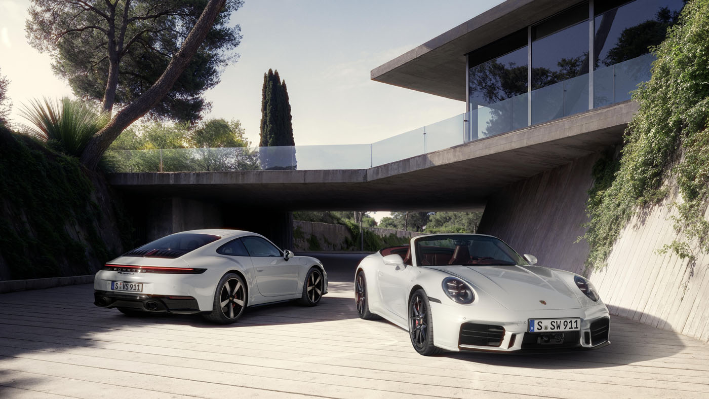 Two silver Porsche 911 sports cars, one coupe and one convertible, parked outside a modern house surrounded by greenery—a perfect showcase for Porsche’s 2025 reveals.
