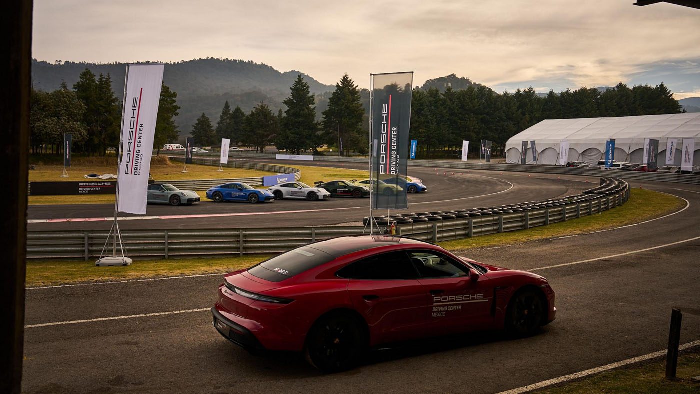 A red Porsche is parked on a racetrack near a curve, with several cars lined up on the other side and banners and trees in the background.