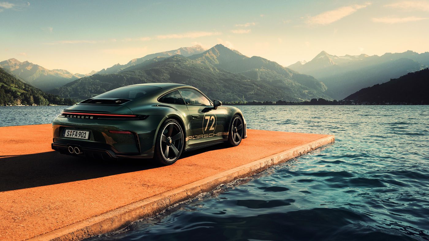 A green Porsche 911 GT3 sports car with the number 72 is parked on a concrete platform beside a lake, mountains rising behind—a striking tribute for F. A. Porsche’s 90th birthday under a partly cloudy sky.