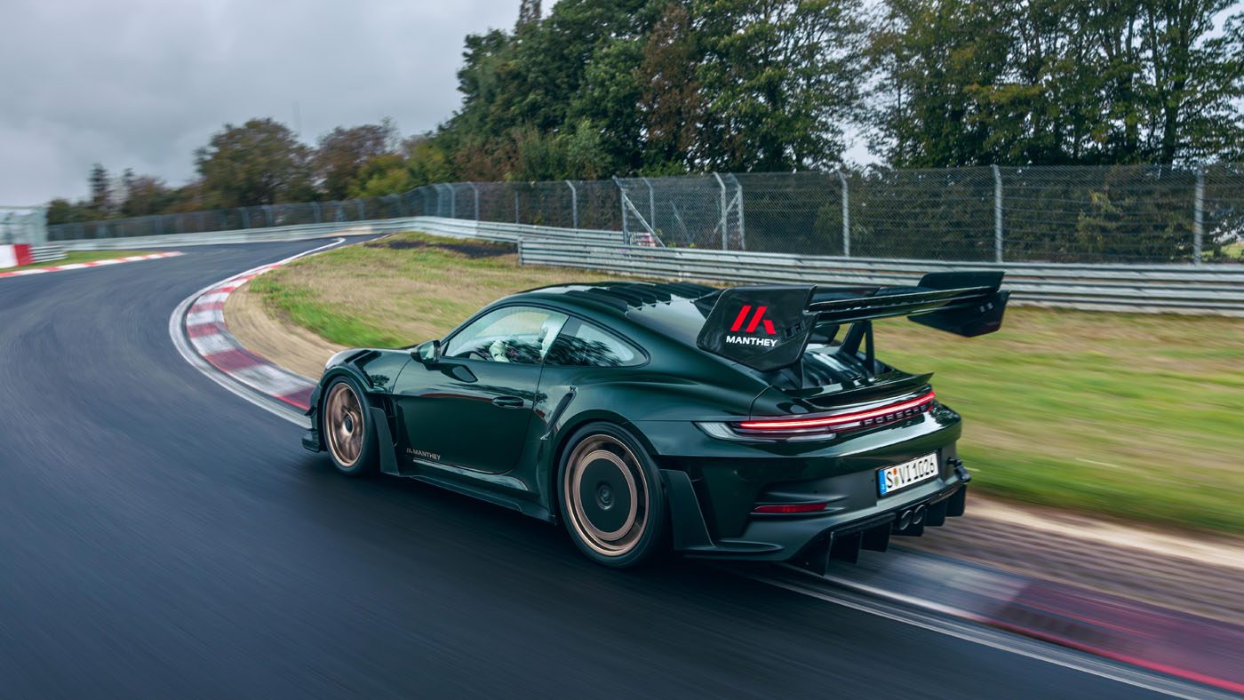 A black Porsche 911 GT3 RS, enhanced with a new Manthey Racing kit, zooms along a curved track surrounded by trees and guardrails, its large rear spoiler slicing effortlessly through the air.