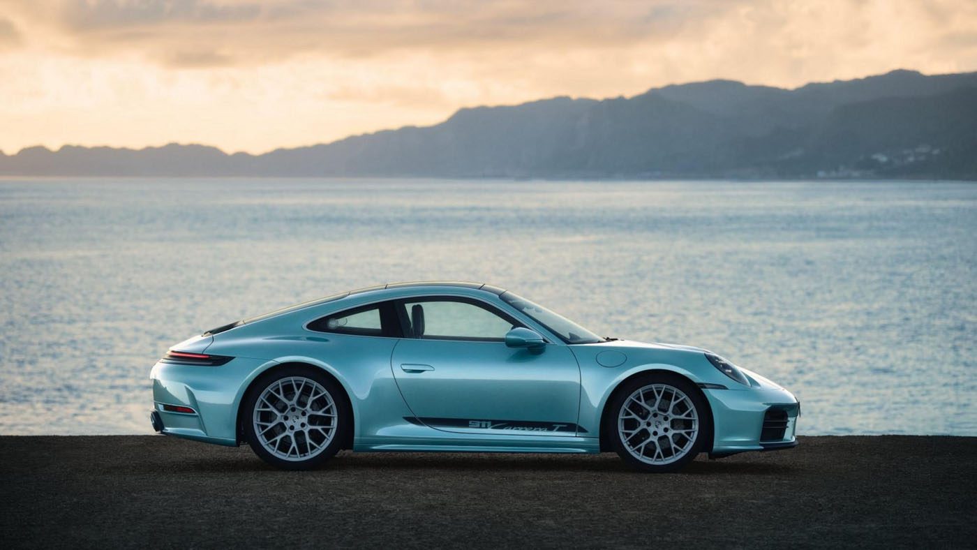A silver Sonderwunsch 911 Carrera T is parked beside a body of water, with mountains rising in the background under a cloudy sky—a stunning Porsche scene reminiscent of Formosa’s scenic beauty.