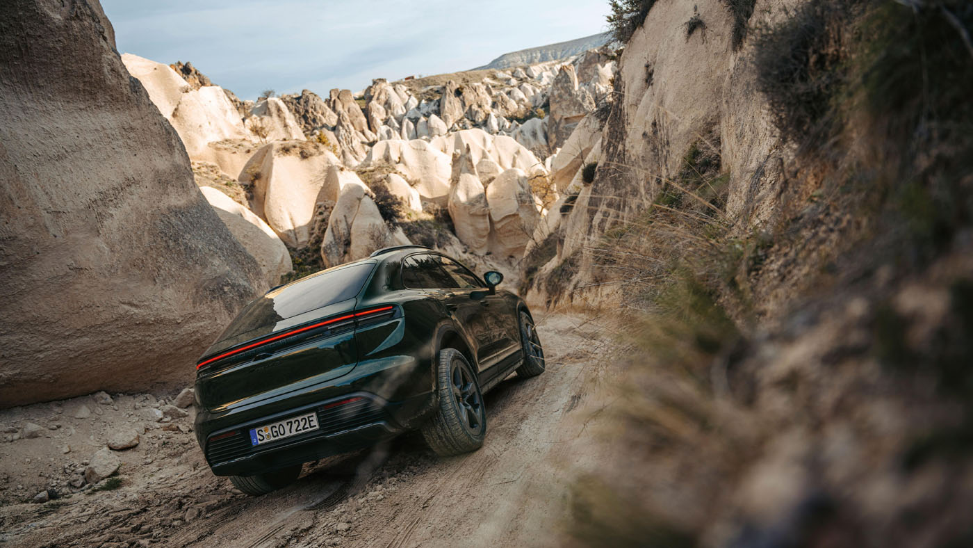 A Porsche Macan Electric navigates a narrow, rocky dirt road in Cappadocia, Turkey, surrounded by large beige rock formations and sparse vegetation.
