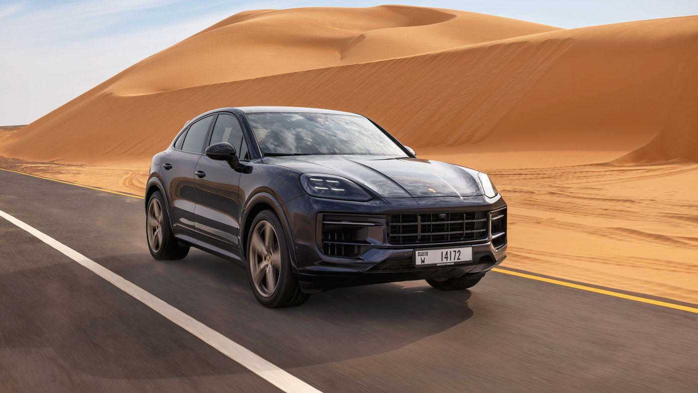 A dark blue Cayenne S Coupe cruises along a paved road, navigating the desert landscape with large sand dunes in the background, as part of an exhilarating Porsche Road Trip from Dubai to Abu Dhabi.