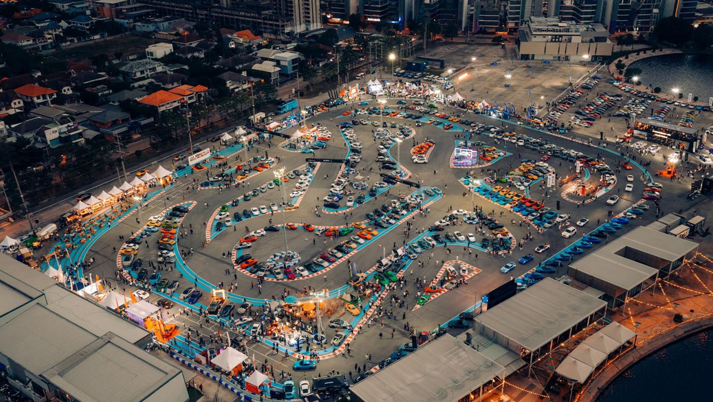 Aerial view of a large outdoor go-kart racing track surrounded by parked cars, spectators, and city buildings at dusk.