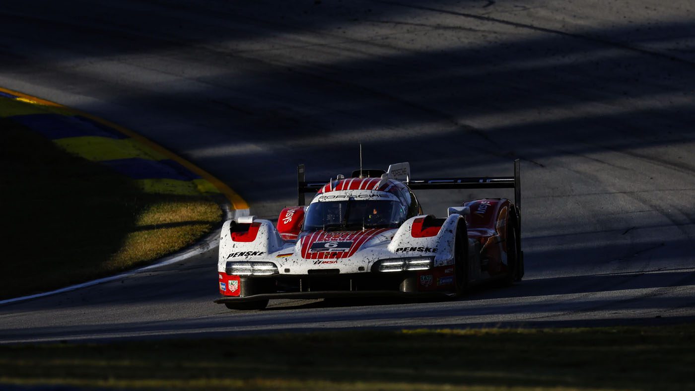 A red and white Porsche race car with visible dirt on the front drives on a sunlit section of the IMSA WeatherTech SportsCar racetrack, casting shadows in the background, embodying the thrill of 2025 motorsport.