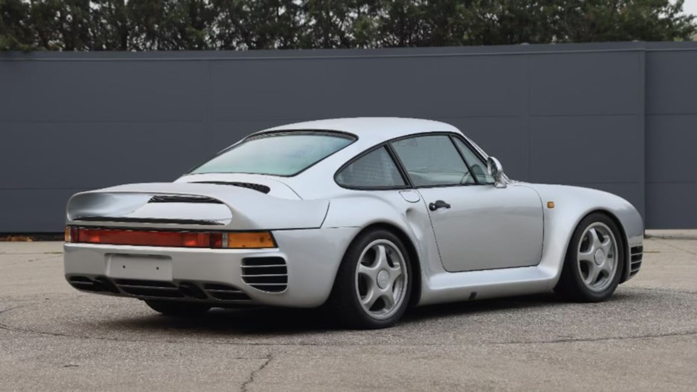 A silver sports car, viewed from the rear left, is parked on a paved surface in front of a gray wall with trees in the background—an elegant example of Porsche Classic Restoration for the Area South Central Restoration Challenge.