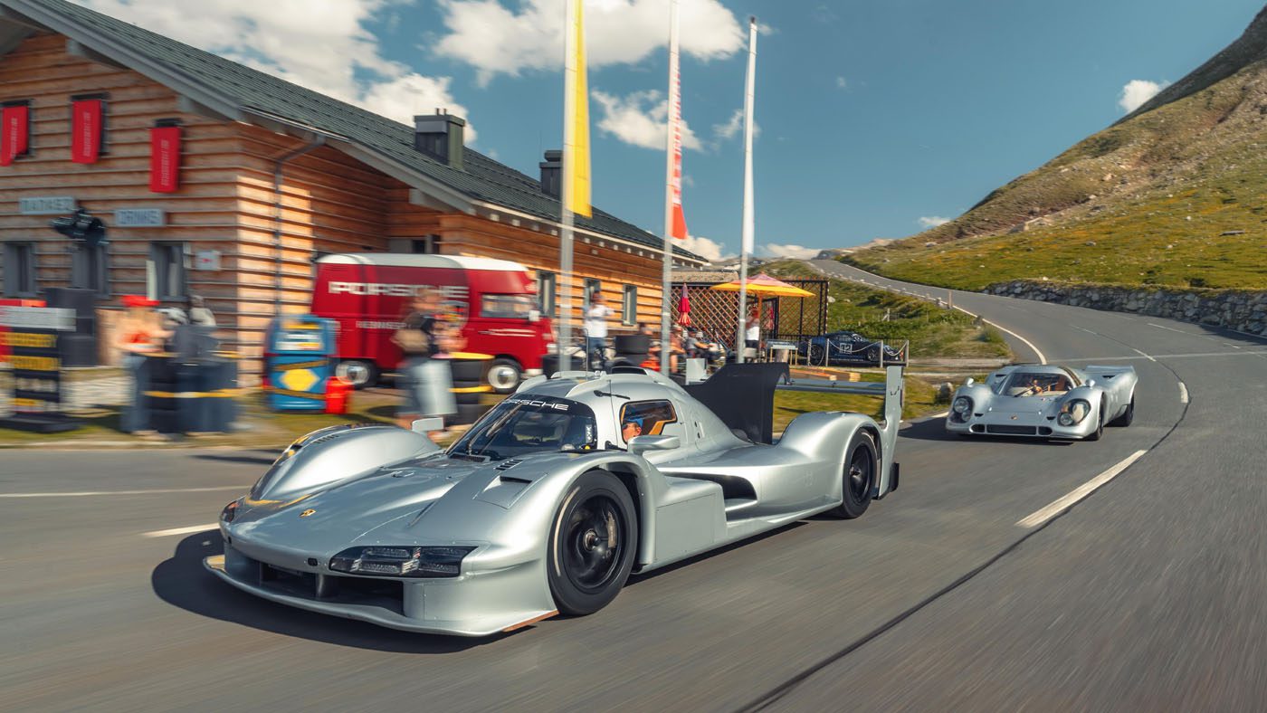 Two silver Porsche 963 RSP race cars drive on a mountain road near the FAT Mankei wooden building, where people stand outside under a sunny sky.