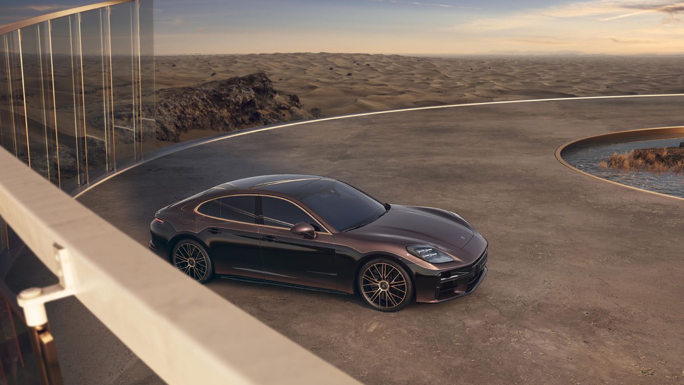 A dark-colored luxury sedan is parked on a circular concrete platform with a glass railing, overlooking a desert landscape at sunset.