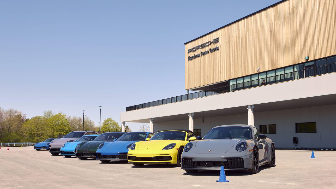 A lineup of six Porsche sports cars in various colors parked outside the Porsche Experience Center Toronto on a sunny day.