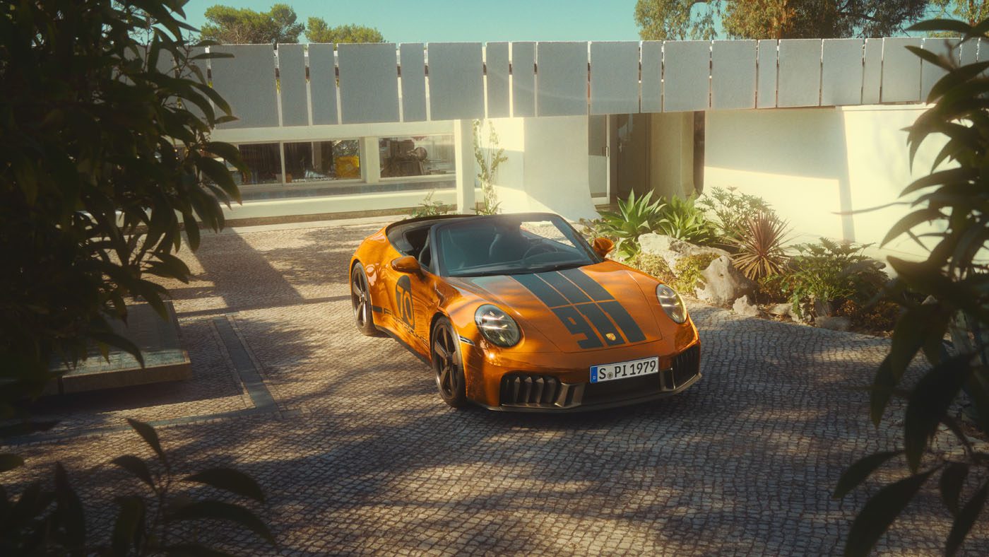 A limited edition orange Porsche 911 convertible with black racing stripes is parked on a paved driveway in front of a modern white house, surrounded by greenery.