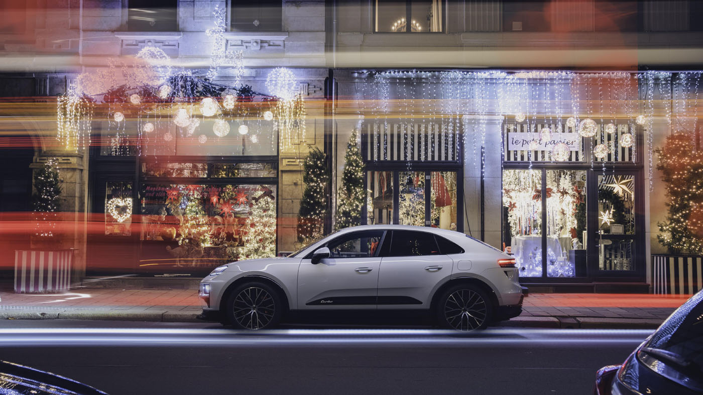 A white Porsche Macan Electric SUV is parked on a city street at night in front of shops decked with Christmas lights and ornaments, with blurred light trails from passing traffic, capturing the spirit of a Porsche Christmas.