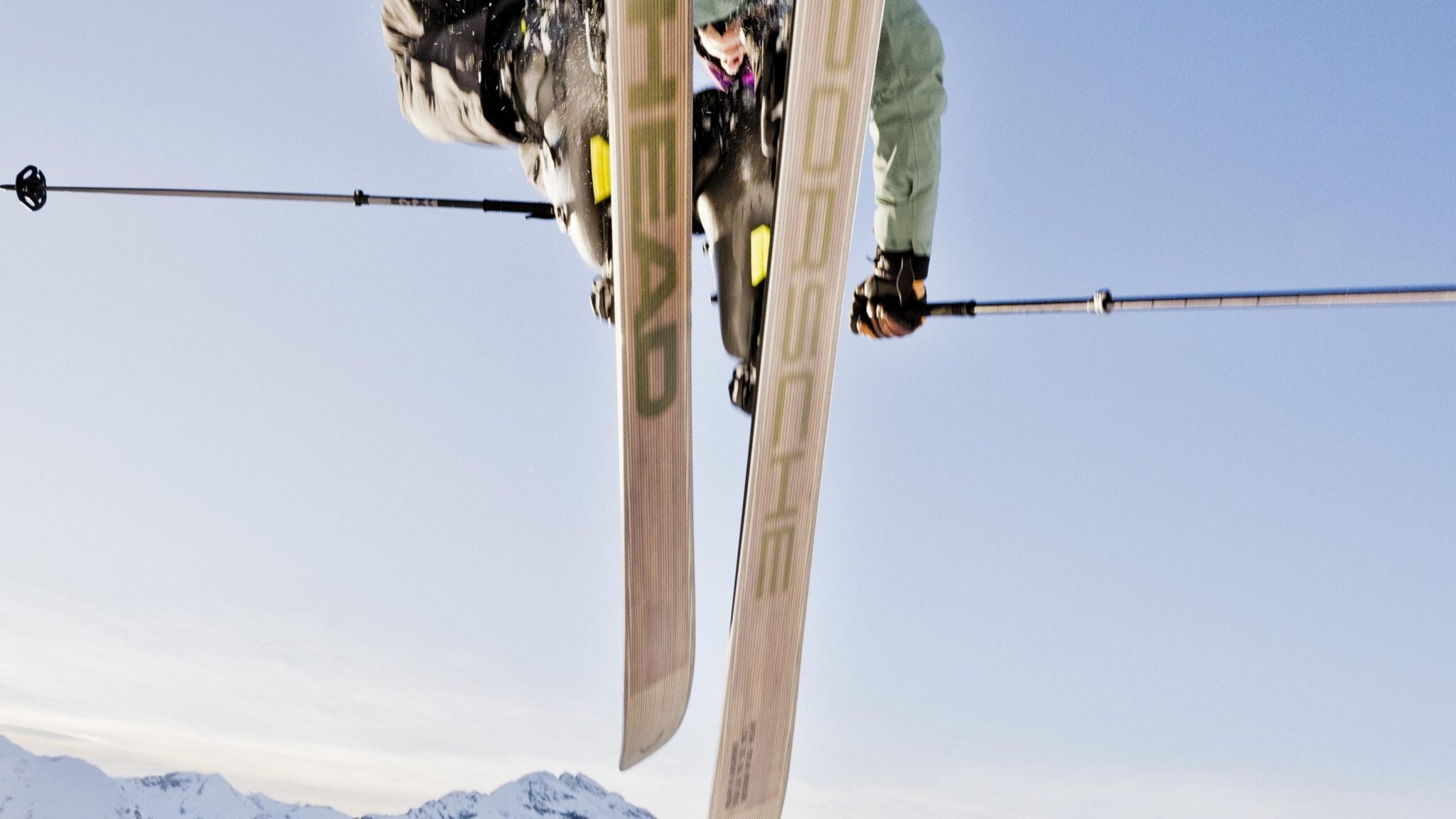 A skier executes a mid-air trick against a clear blue sky, with snow-covered mountains in the background, showcasing precision akin to sports car engineering.