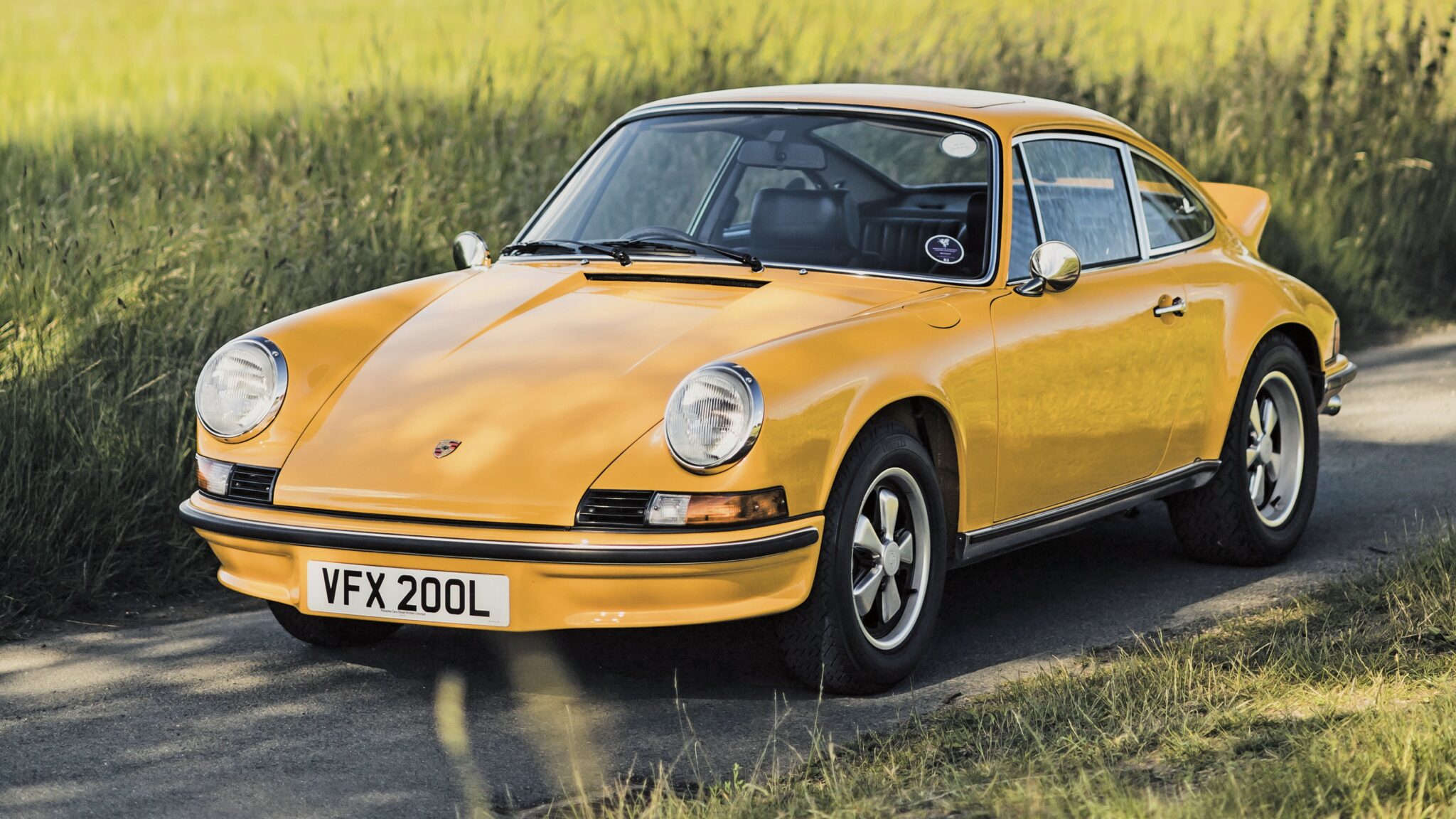 A yellow Porsche 911 Carrera RS 2.7 rests elegantly on a rural road, grass lining both sides. It’s a sight even a classic car expert like Simon Kidston would admire, appreciating the timeless beauty and storied design of this vintage masterpiece.