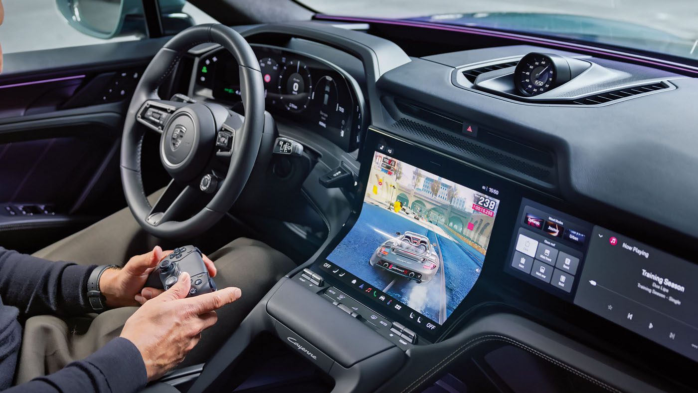 A person sits in a Porsche Cayenne’s driver seat holding a game controller, with a racing video game displayed on the car’s central touchscreen, blending immersive driver experience with digital entertainment.