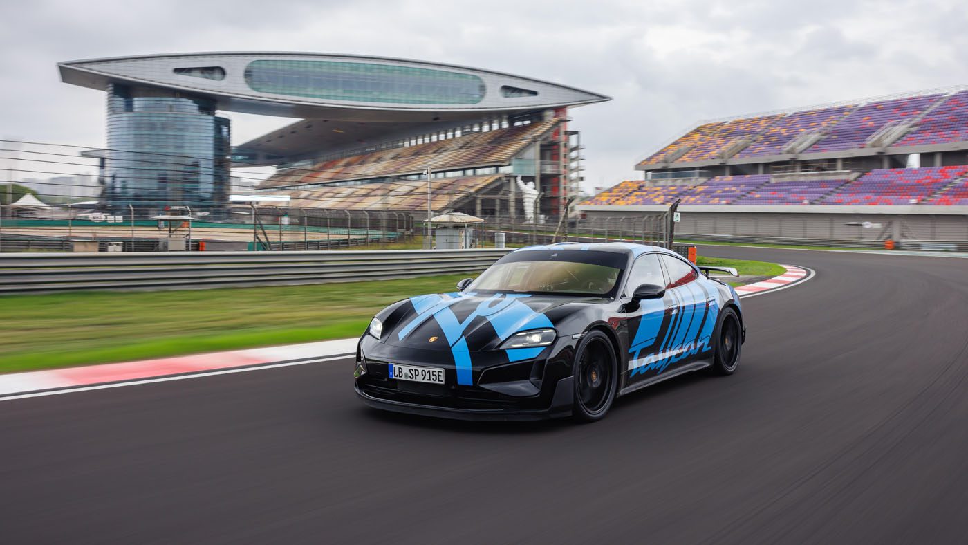 A sleek, black and blue Turbo GT sports car speeds around the racetrack at Shanghai International Circuit, with grandstands and a modern building in the background.