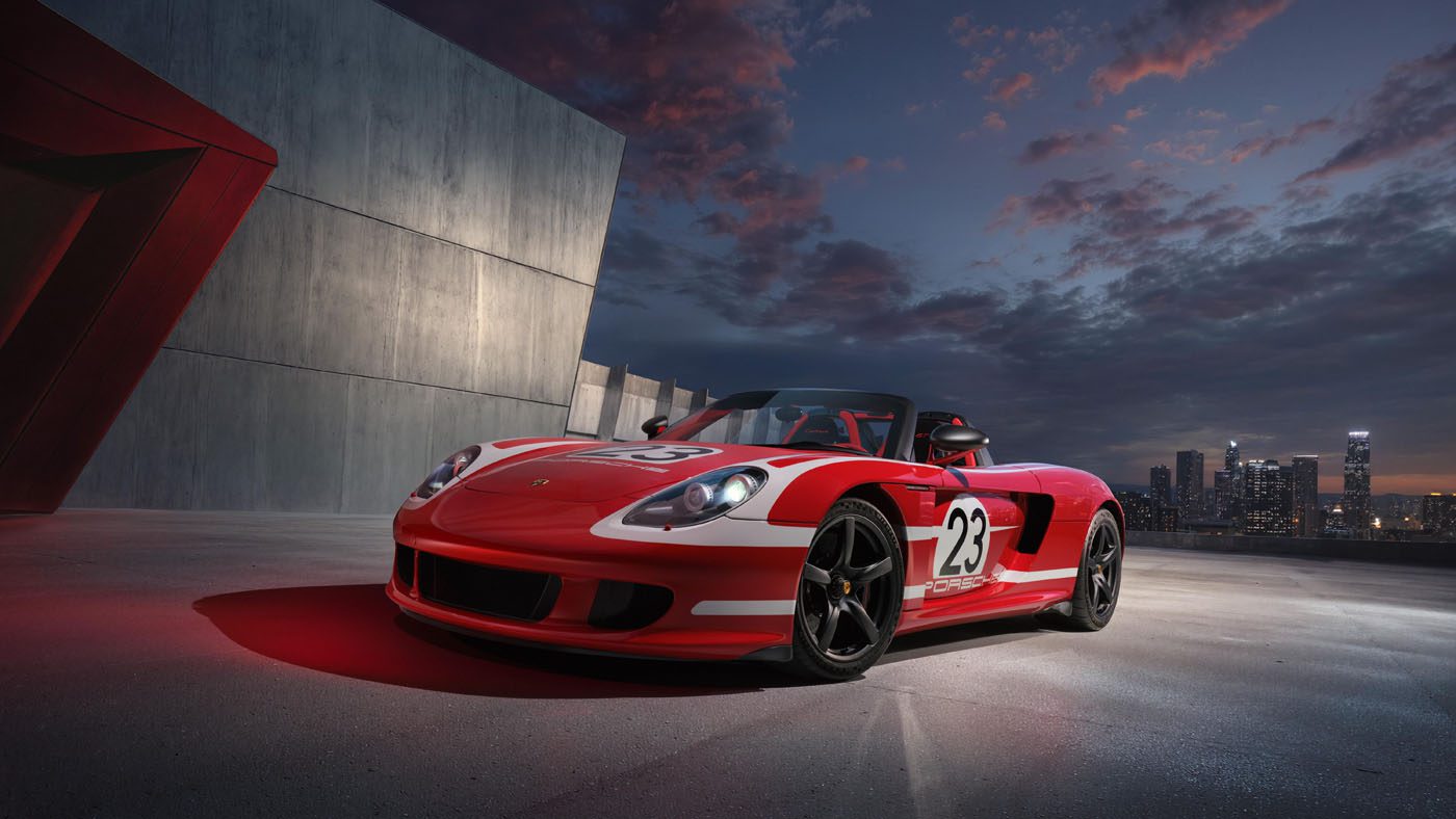A red and white Porsche Salzburg Carrera GT with the number 23 on its side is parked on a rooftop at dusk, framed by the city skyline in the background.