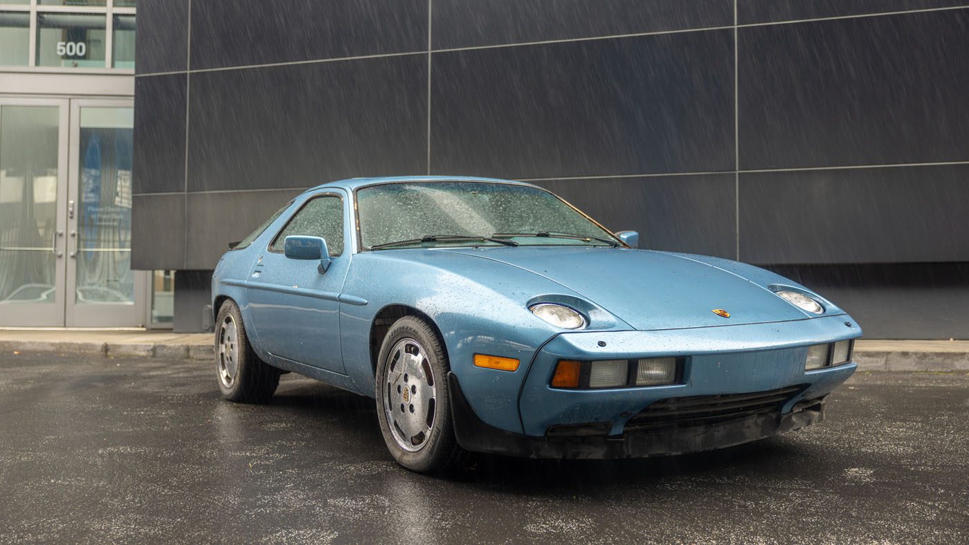 A blue Porsche 928 sports car, part of the Porsche Classic Restoration Challenge, is parked on wet pavement in front of a modern building on a rainy day.