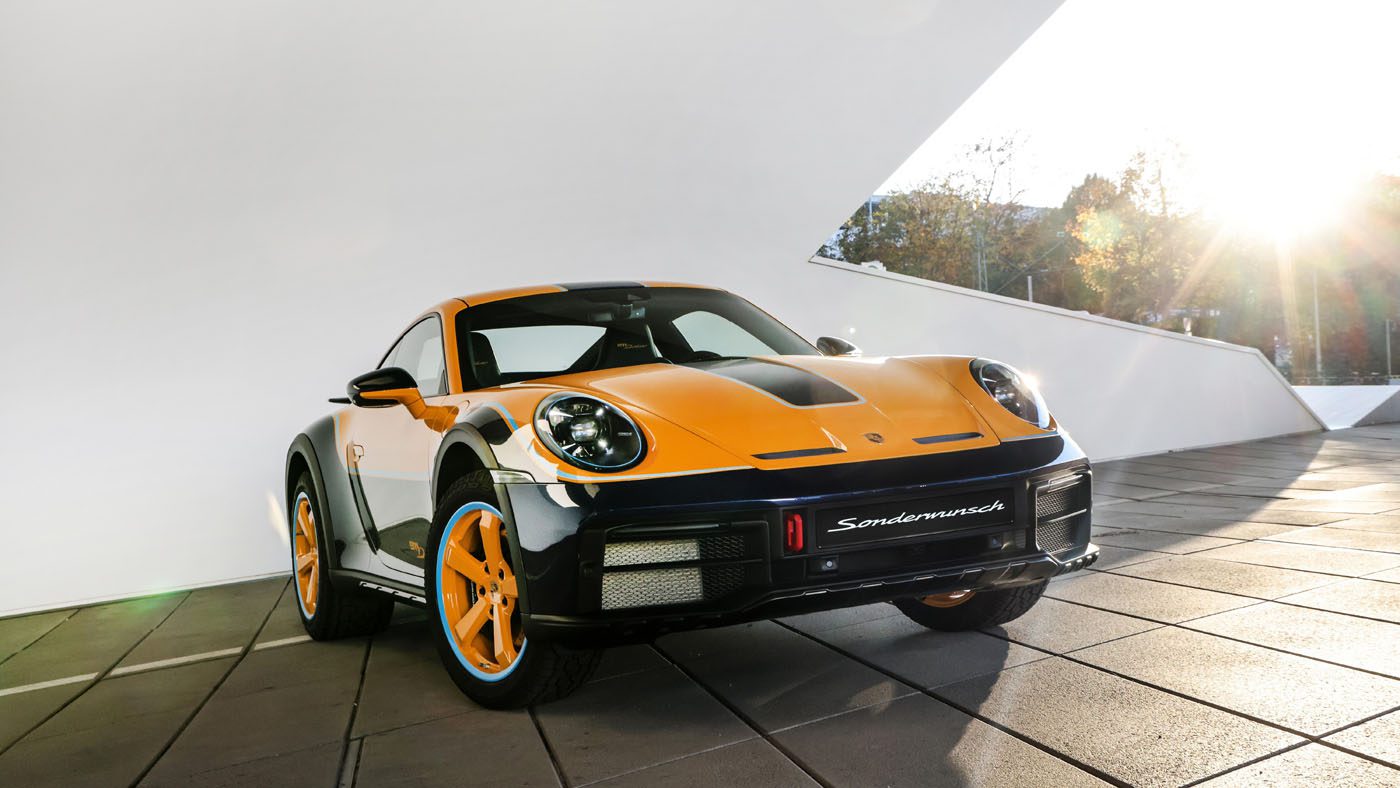 The one-off black and orange Porsche 911 Dakar is elegantly parked on a tiled surface, with a sleek modern building rising majestically in the background.