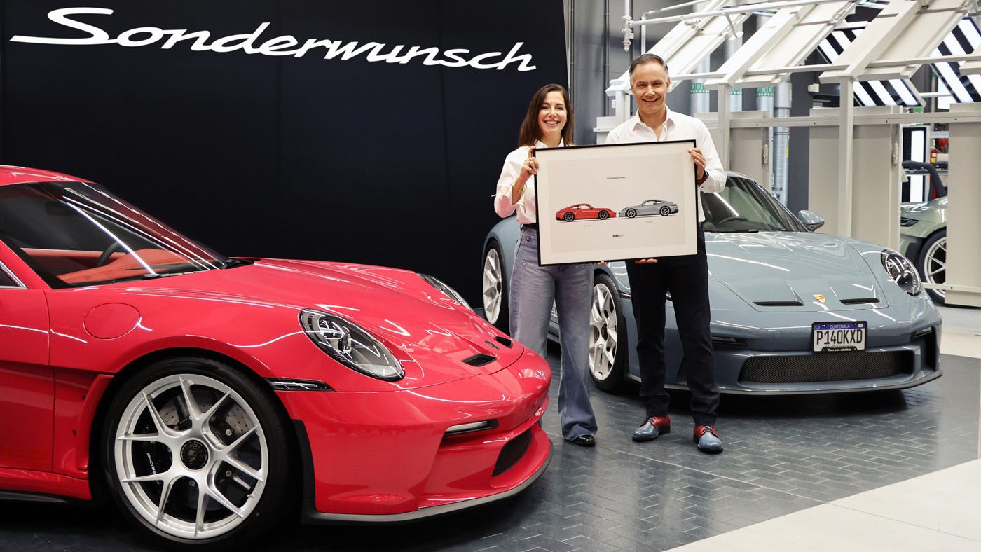A man and woman stand between a red and a grey Porsche 911 S/T, holding a framed sketch of the two cars, in front of a sign that reads "Sonderwunsch.