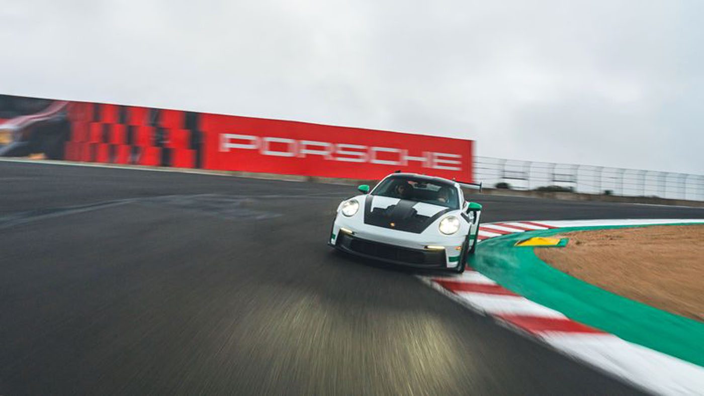 A white Porsche Official Car takes a sharp turn on WeatherTech Raceway Laguna Seca, with a red Porsche sign in the background.
