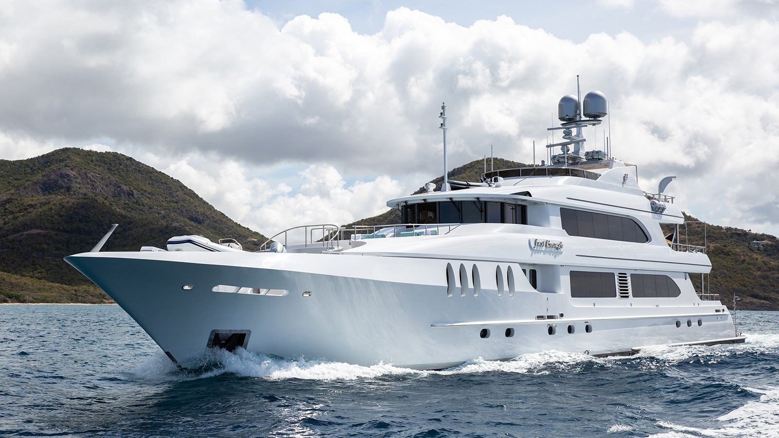A large white luxury yacht cruises on blue ocean water with green hills and a cloudy sky in the background, offering a true maritime experience.