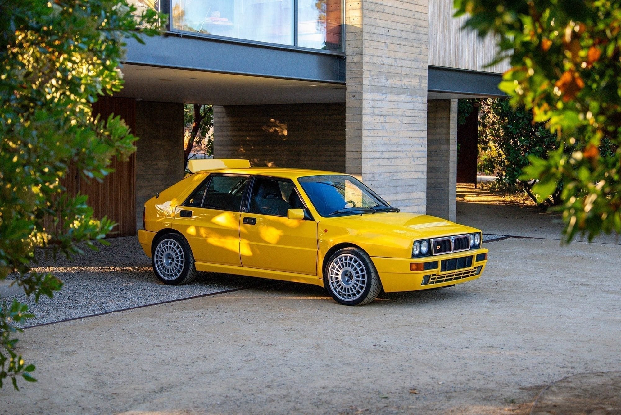 A yellow Lancia Delta Integrale hatchback is parked on a driveway in front of a modern house, surrounded by trees and sunlight—a true Petrolicious moment.