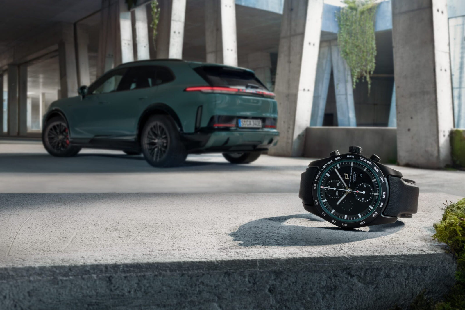 A wristwatch from ICustom Timepieces with a black strap rests on a concrete surface in the foreground, while a green SUV from the Porsche Design Cayenne Models is parked in the background under a modern structure.