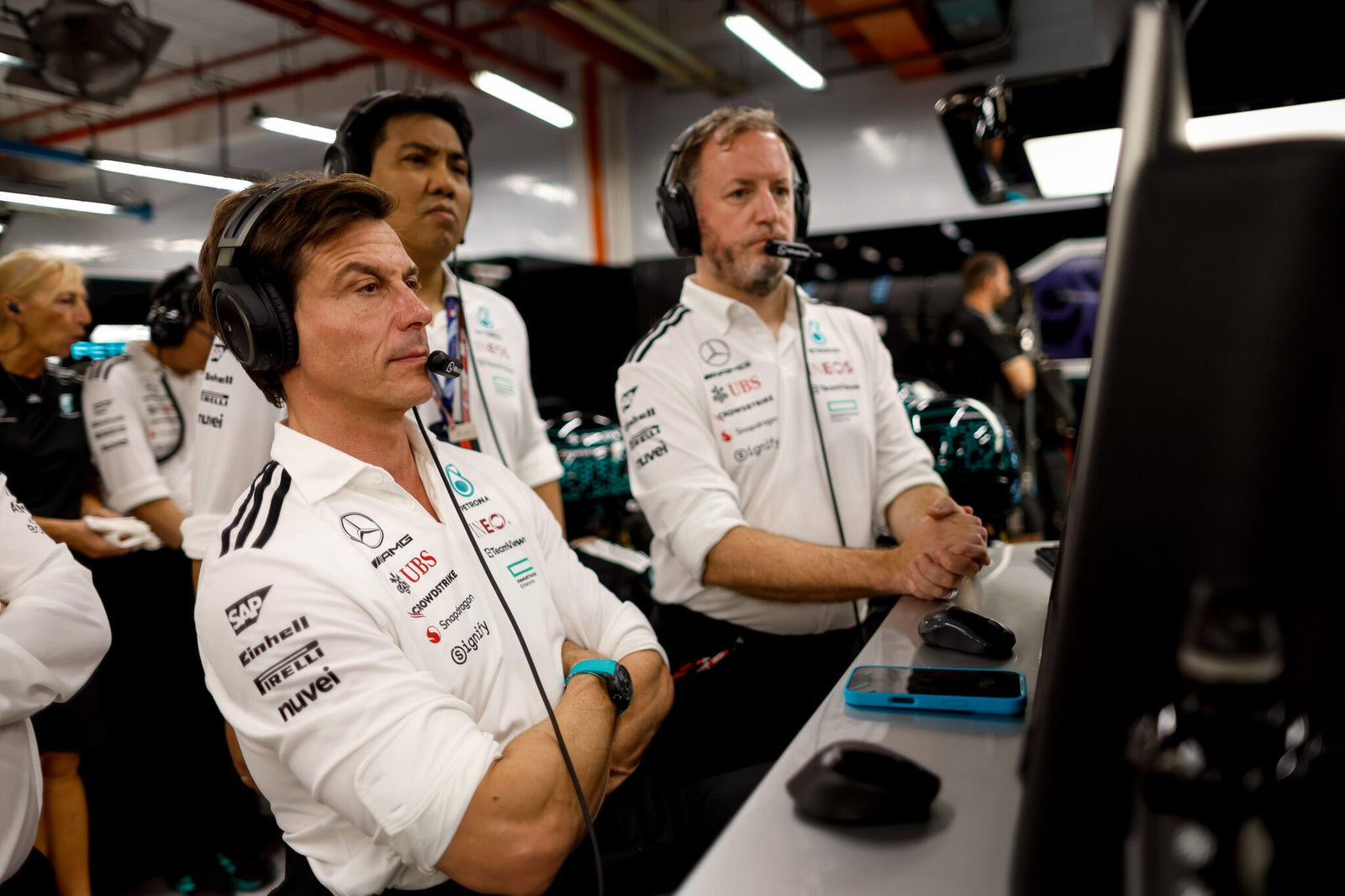 Three men in Mercedes-AMG Petronas team uniforms wearing headsets watch monitors intently inside a Formula 1 garage, with Toto Wolff focused on strategy alongside his team.