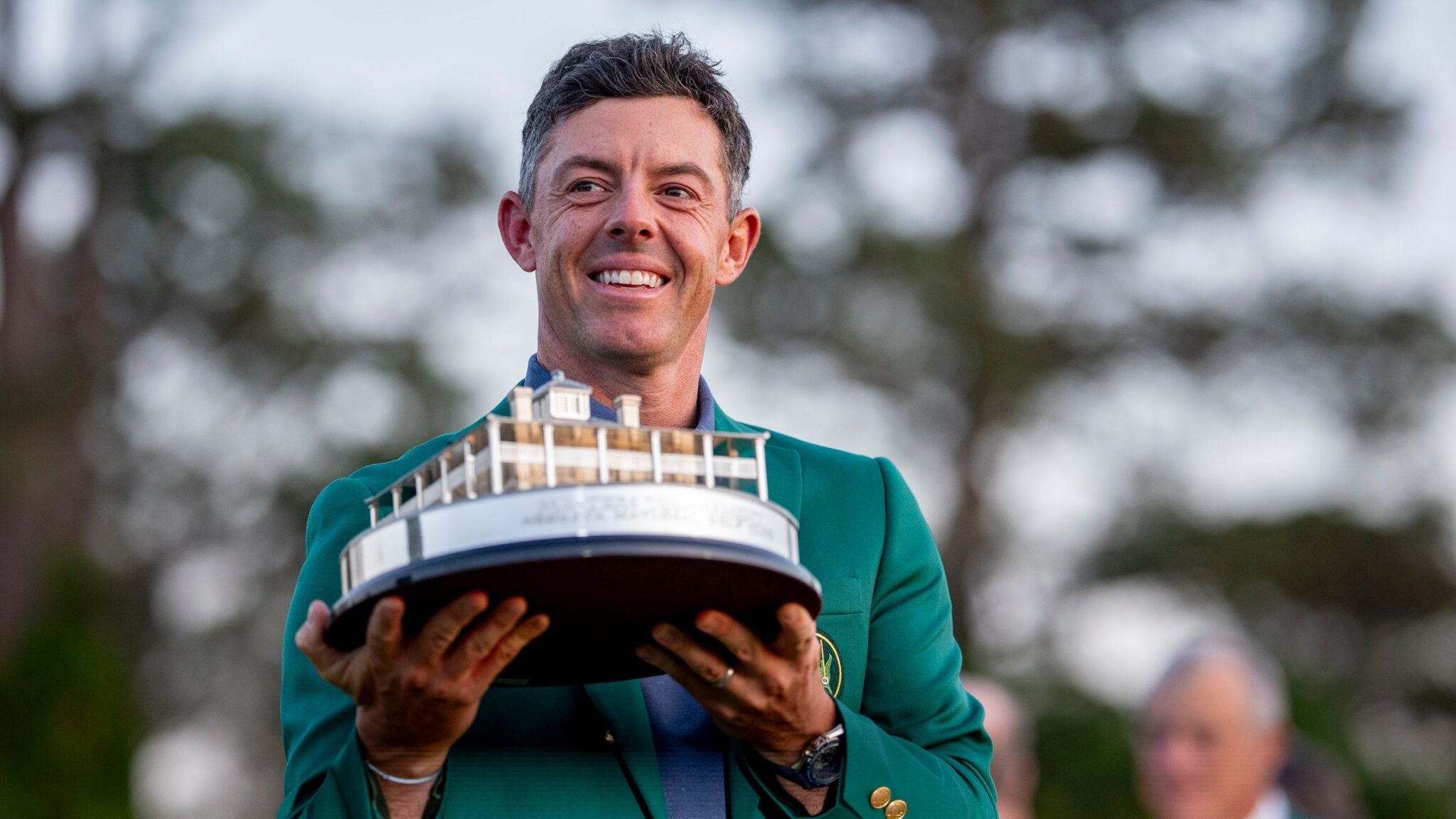 A person in a green jacket holds a trophy shaped like a building, reminiscent of Rory McIlroy's Masters win, standing outdoors with trees in the background.