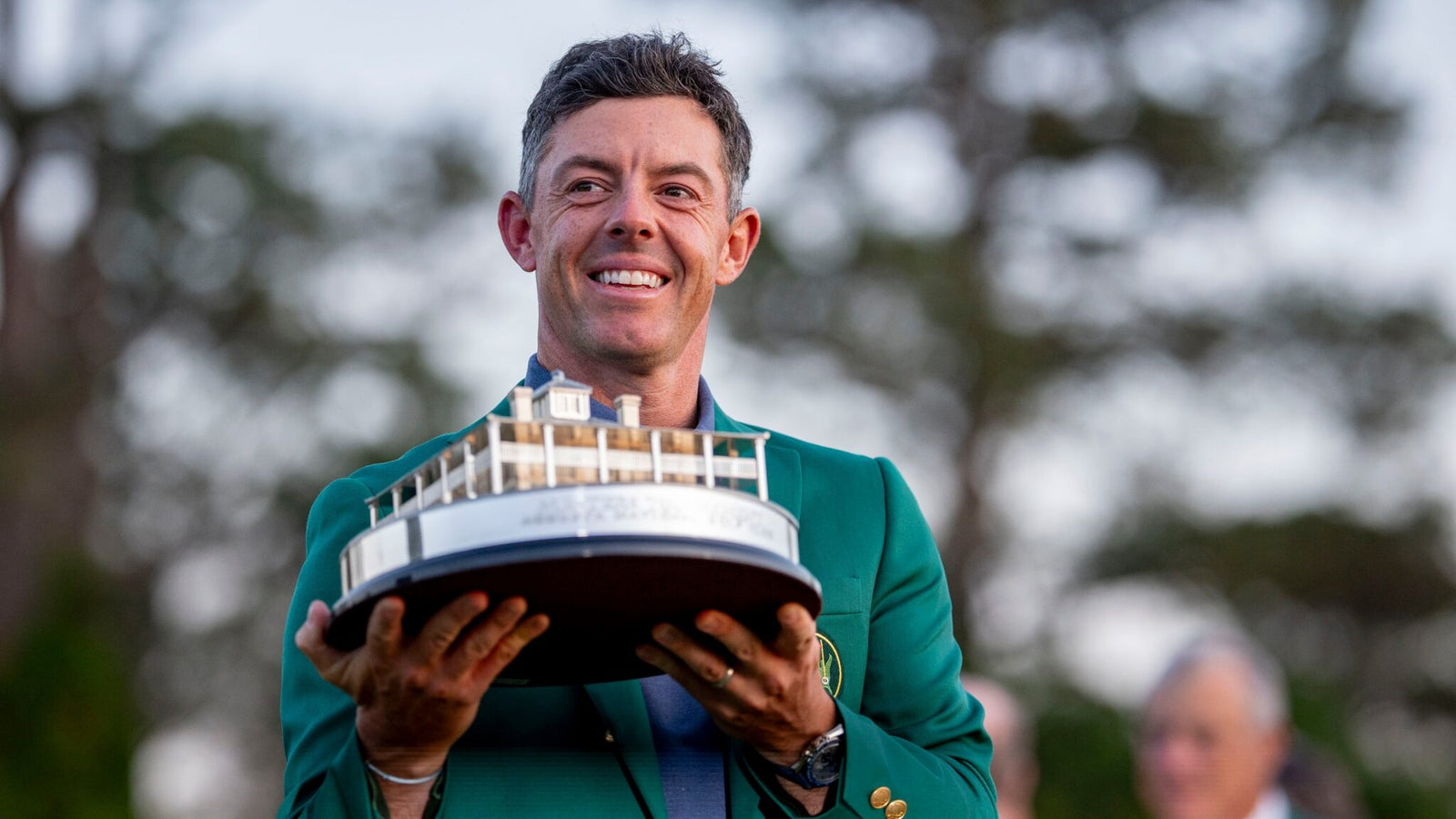 A person in a green jacket holds a trophy shaped like a building, reminiscent of Rory McIlroy's Masters win, standing outdoors with trees in the background.