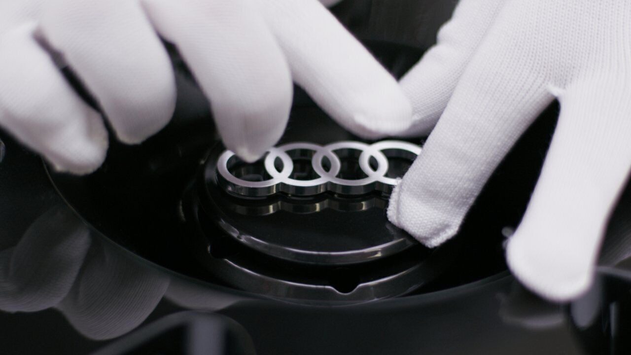 A person wearing white gloves holds a car part featuring the silver Audi logo with four interlinked rings, as seen in a making-of video for a concept car.