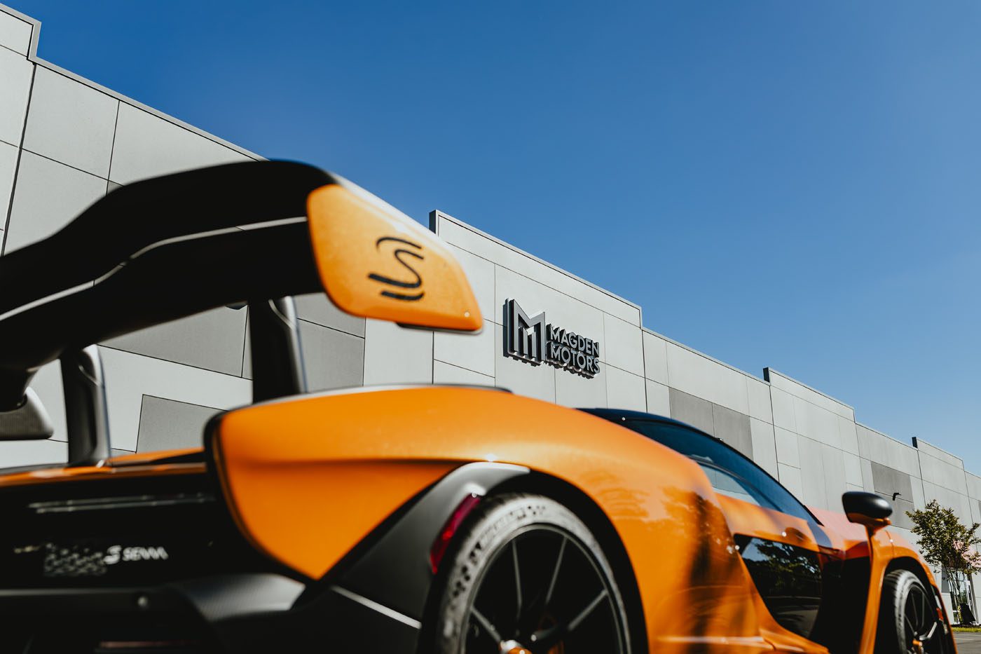 An orange sports car with a prominent rear spoiler is parked in front of a building adorned with "Exclusive Dealer" signage under a clear blue sky, capturing the essence of Jessup, MD.