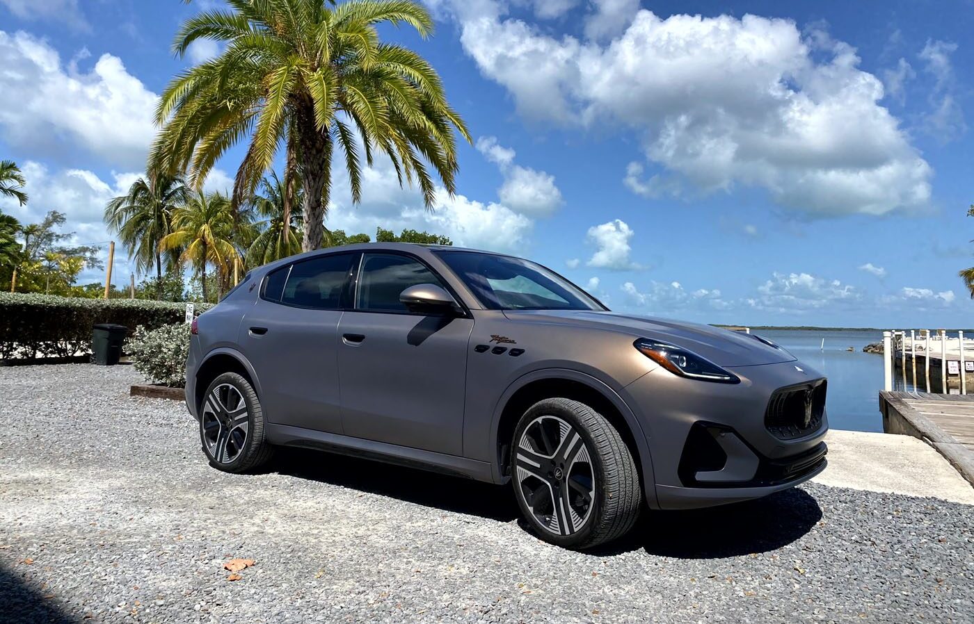 A matte gray, all-electric Maserati Grecale Folgore SUV is parked near palm trees by a waterfront in the sunny Florida Keys under a clear sky with scattered clouds.