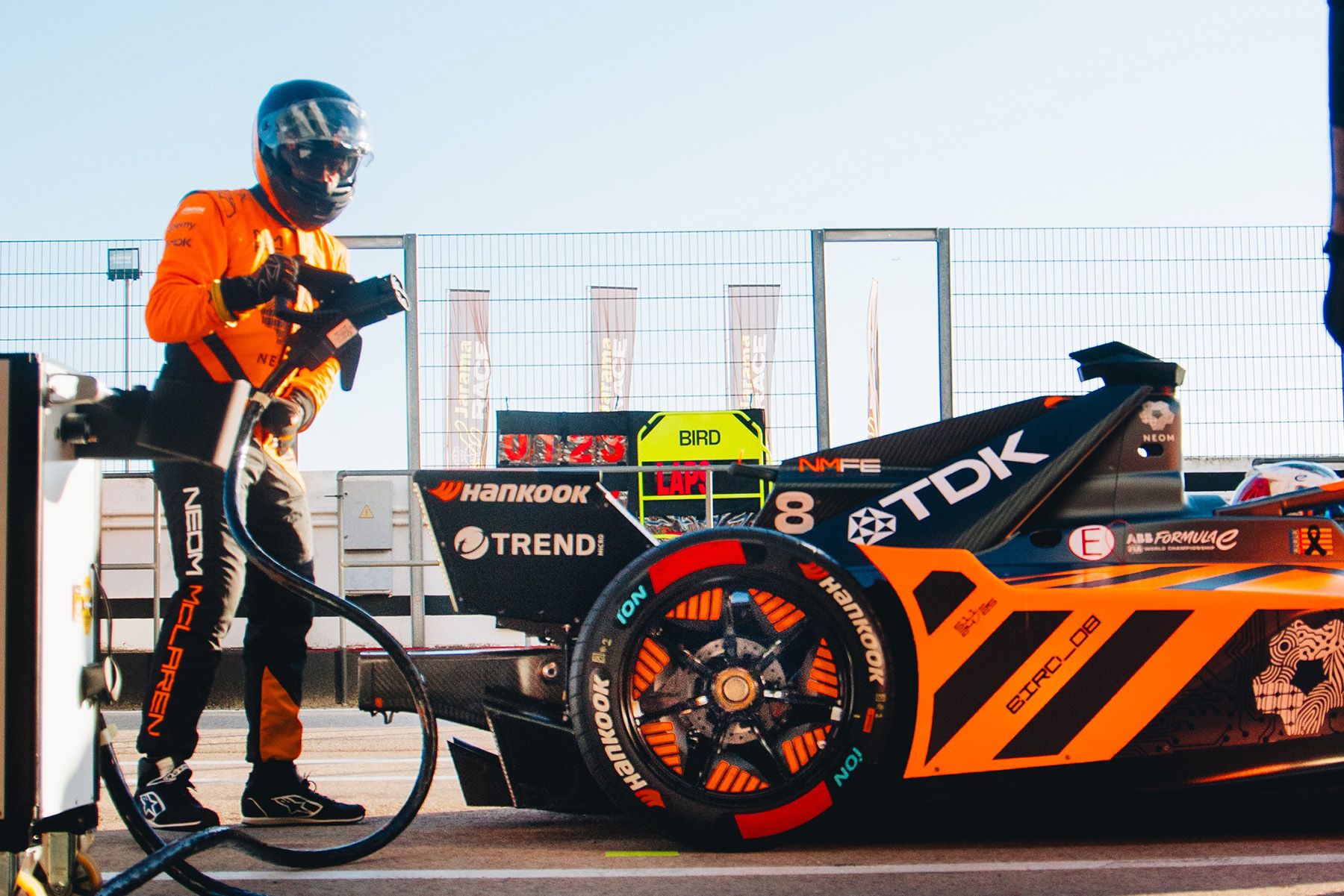 A pit crew member in orange gear prepares to service a McLaren race car marked with the number 8 and various sponsors, parked in a pit lane during the sunny 2024/25 Formula E season.