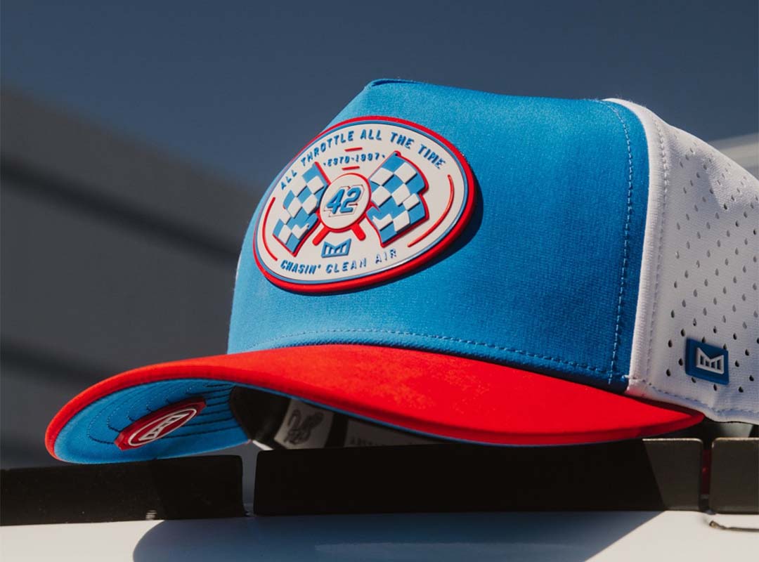 A blue, white, and red Odyssey Hat by melin with a round NASCAR-inspired racing patch on the front, featuring checkered flags and the number 42, resting on a white surface.