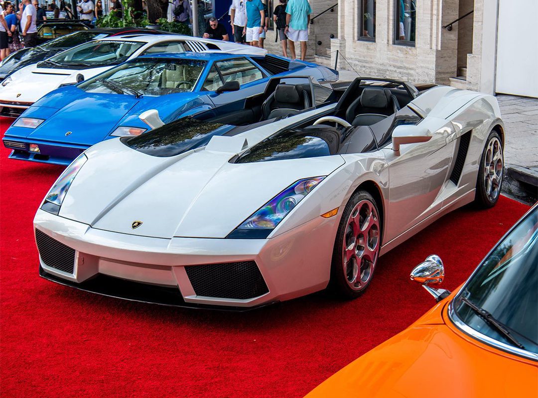 A white convertible sports car gleams on the red carpet amidst other luxury vehicles at the Miami Concours. Elegantly dressed attendees linger in the background beside Moore Private Members’ Club, creating an air of exclusivity and allure.