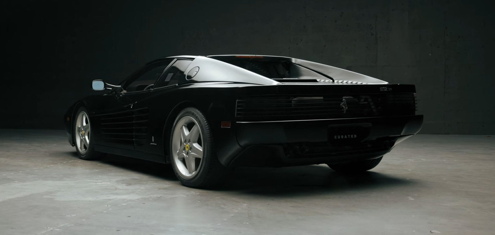 A sleek black Ferrari 512 TR with a low-profile design and angular lines rests on a smooth gray surface in a dimly lit setting, exuding the curated elegance once admired by icons like Michael Jordan.