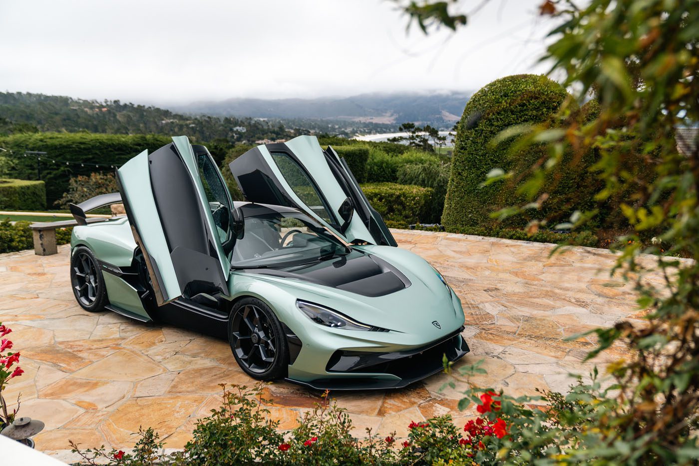 A light green Rimac electric hypercar with its butterfly doors open is parked on a stone driveway surrounded by greenery and flowers, reminiscent of an exclusive Monterey Car Week showcase.