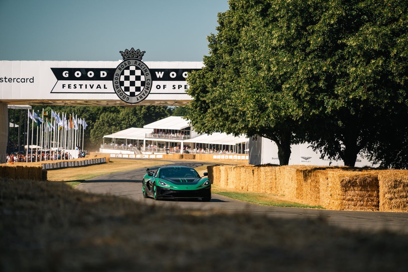 A green sports car drives on a track lined with hay bales at the Goodwood Festival of Speed, with a large event banner and trees in the background.