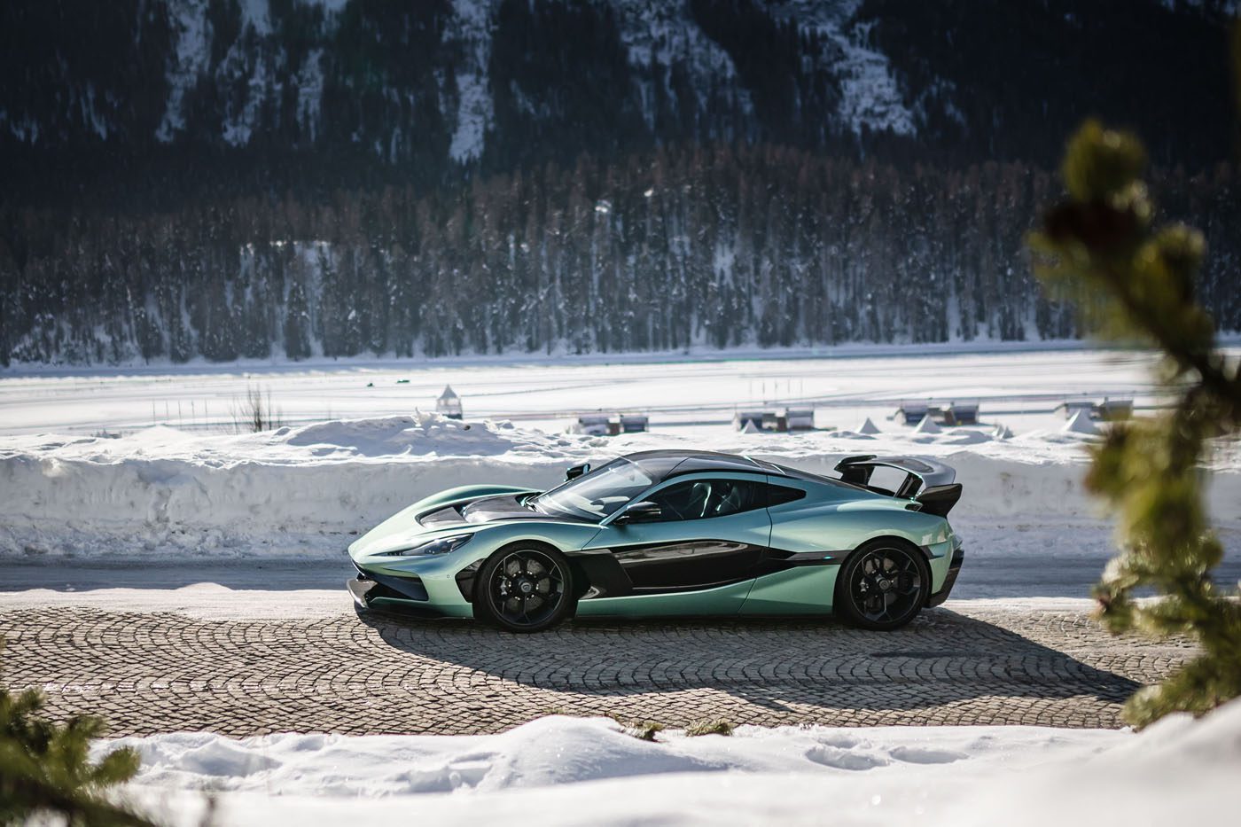 A sleek, metallic green Rimac sports car parked on a cobblestone road in a snowy landscape near I.C.E. St. Moritz, with mountains majestically towering in the background.