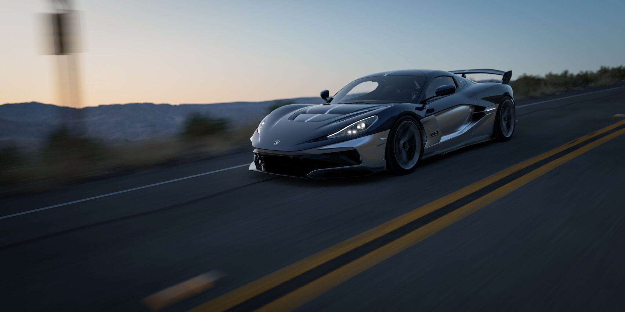 A sleek black Rimac Nevera R hypercar drives at high speed on an empty highway at sunset, with blurred surroundings indicating motion.
