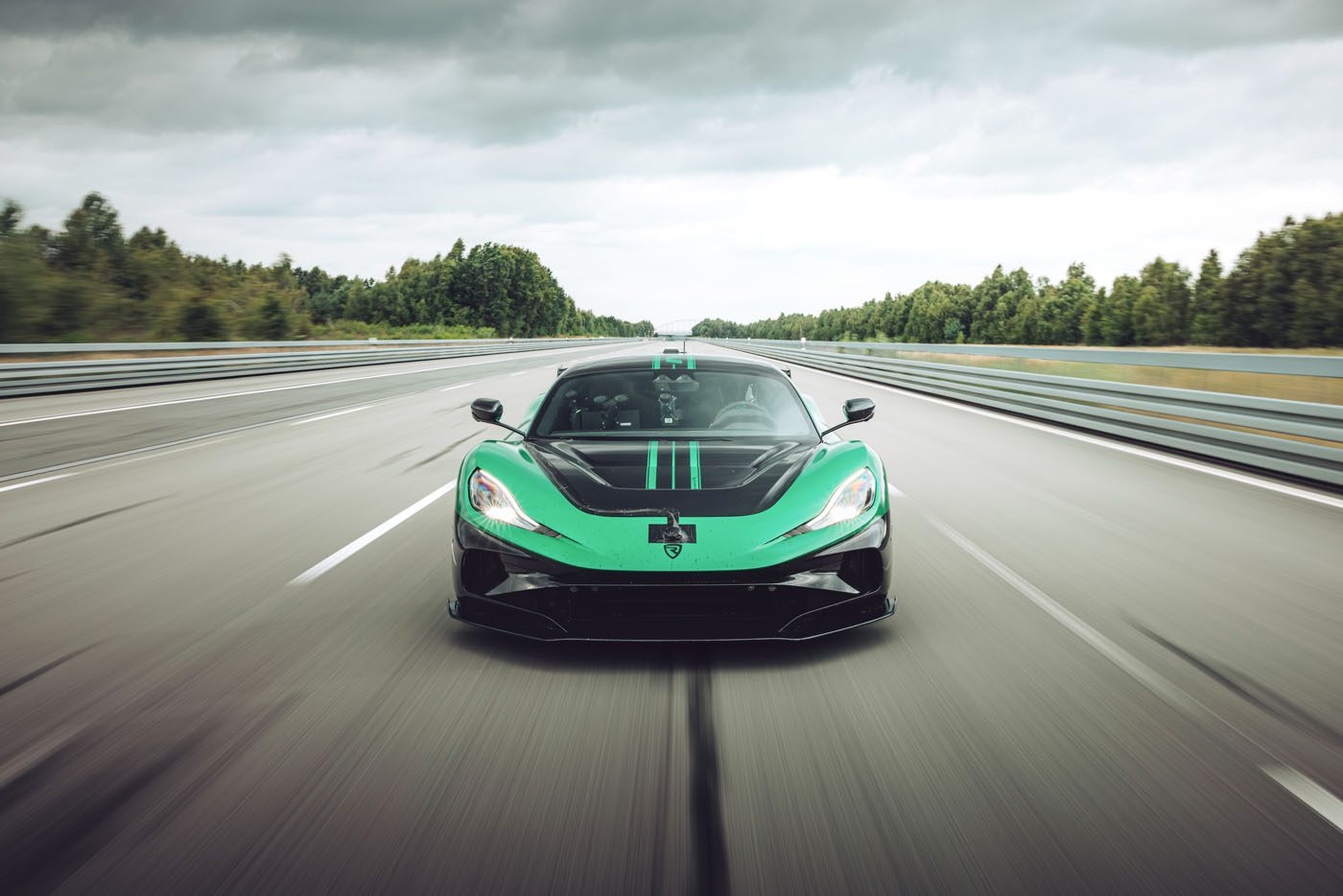 A green and black sports car drives at high speed on an empty highway surrounded by trees under a cloudy sky.