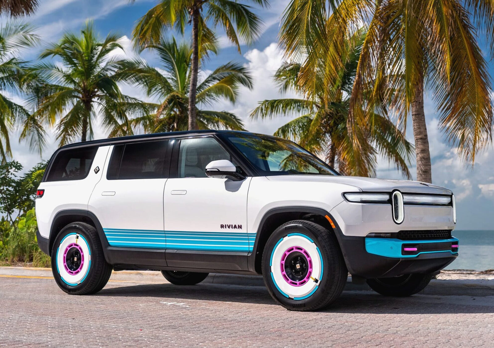 A white Rivian SUV with blue and pink accents is parked on a palm-lined road by the beach—echoing the vibe of the Rivian Opens Brickell Hub With a One-Off Miami-Themed Tri-Motor R1S under a partly cloudy sky.