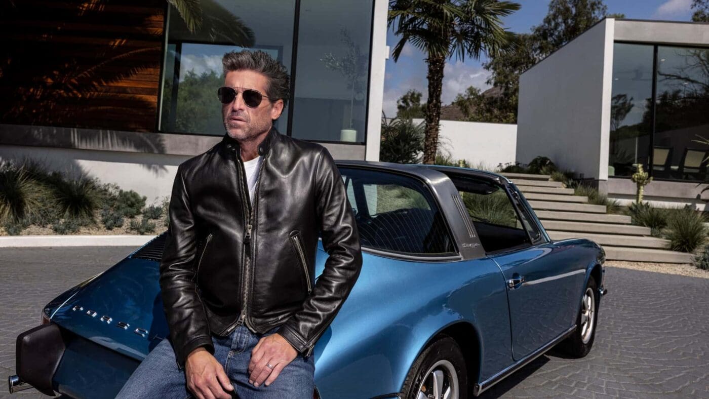 A man wearing sunglasses and a black leather jacket sits on the hood of a blue vintage Porsche, featured in the duPont REGISTRY 2025 Father's Day Gift Guide, parked outside a modern house with palm trees.