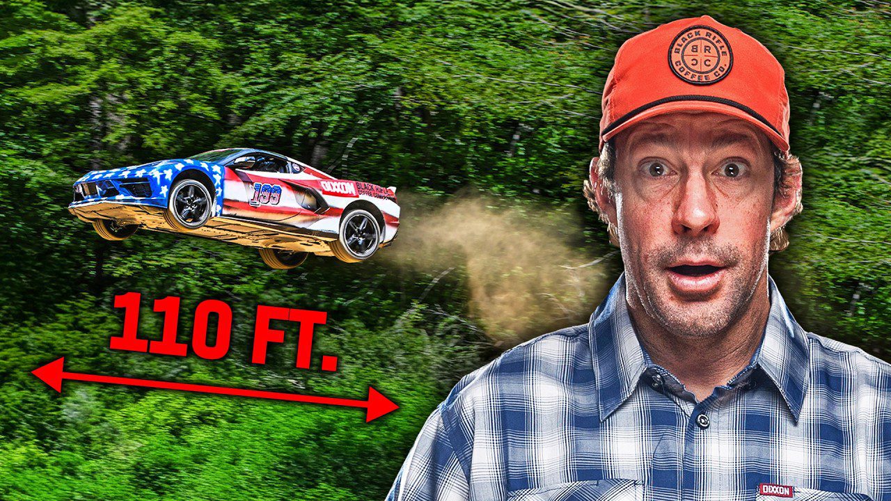 A man in a red cap stands in front of greenery as a C8 Corvette with an American flag design, driven by Travis Pastrana, soars through the air on its longest jump, marked by a "110 FT." label and arrow indicating the distance.