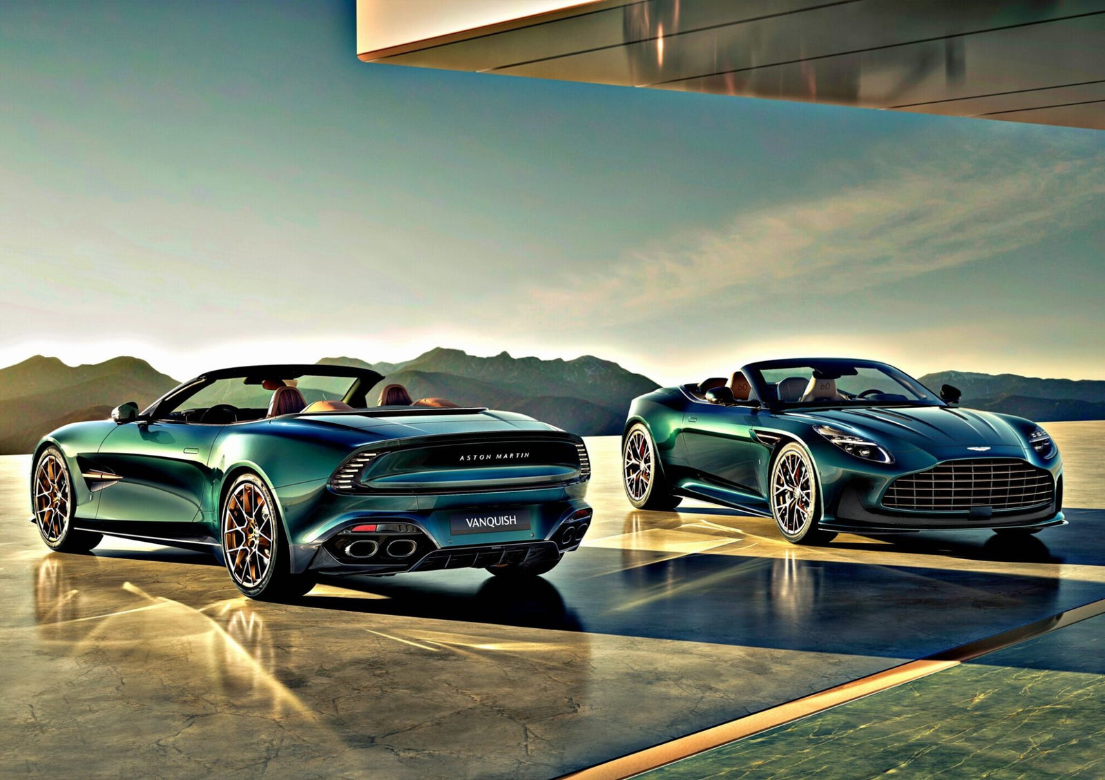 Two green Aston Martin Vanquish convertibles, part of the Q by Aston Martin Marks 60 Years of Volante with Ultra-Exclusive Anniversary Editions, are parked on a sleek terrace with mountains behind and sunlight glinting off their curves.