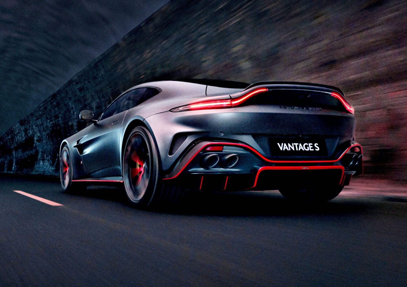 Rear view of a 2026 Aston Martin Vantage S in silver and red speeding along a road, with a stone wall lining the side.