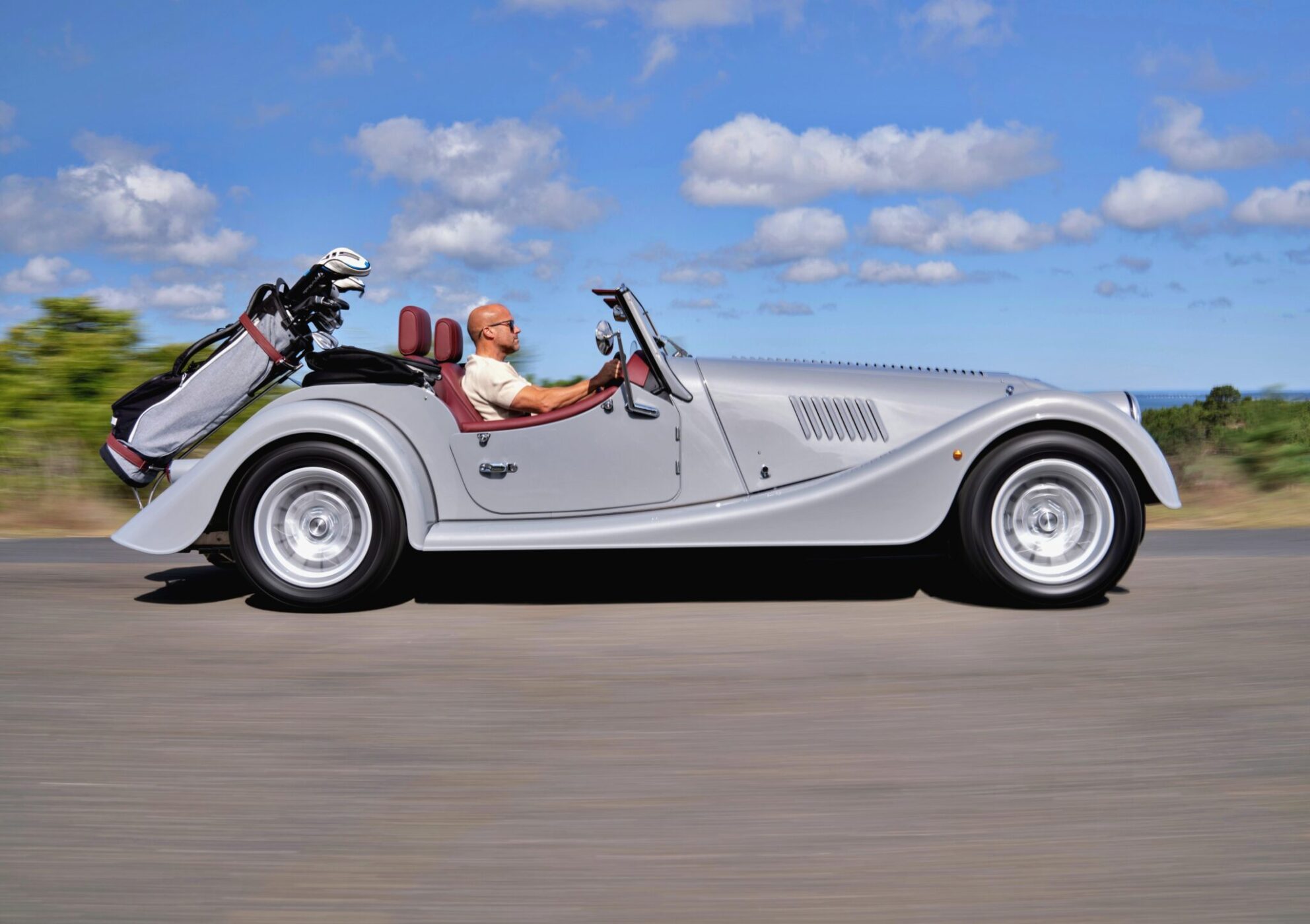 A man drives a silver Morgan Plus Four convertible with a set of golf clubs in the back, cruising down an open Long Island road under a partly cloudy sky.