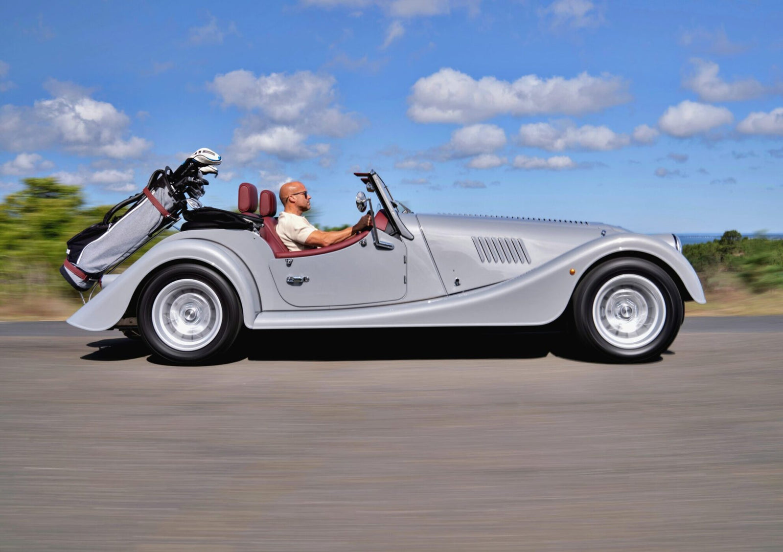 A man drives a silver Morgan Plus Four convertible with a set of golf clubs in the back, cruising down an open Long Island road under a partly cloudy sky.